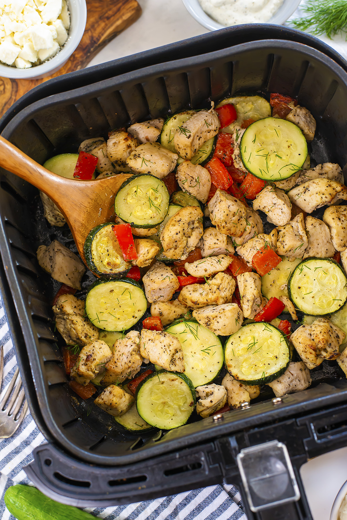 A wooden spoon in an air fryer basket filled with Greek chicken, zucchini and red peppers.