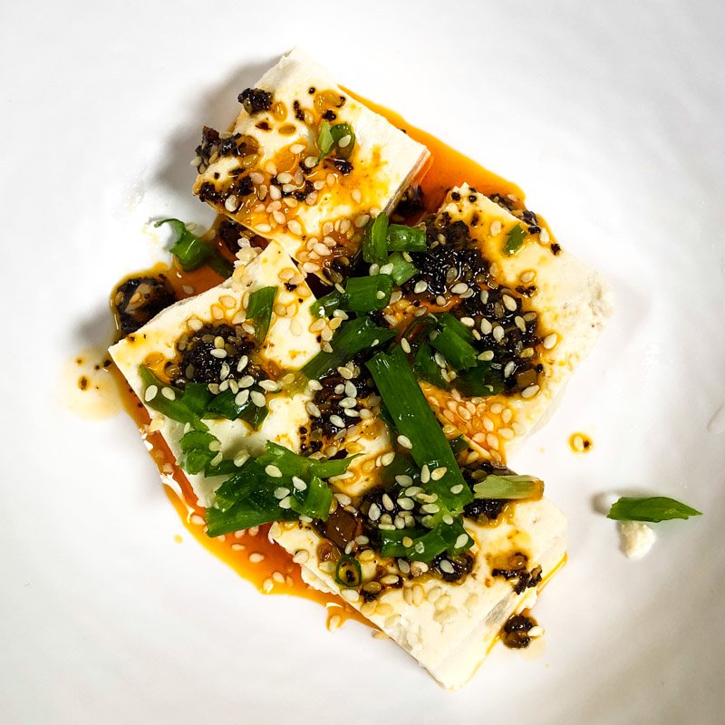 Tofu in Stücken mit Chiliöl und gehacktem Schnittlauch auf einem Teller.