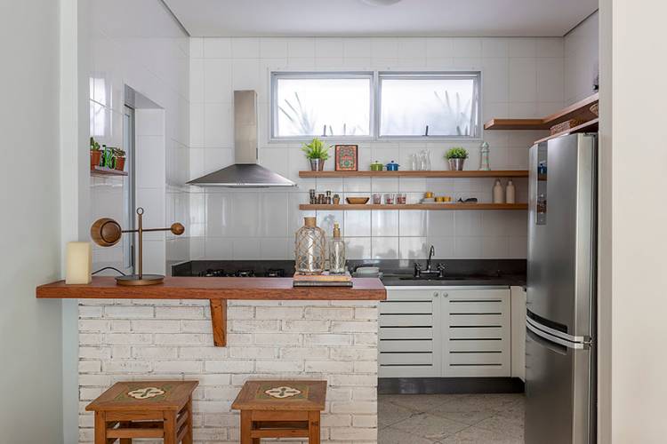 Kitchen with American counter, exposed brick, wooden shelves and bench, white cabinets, refrigerator, hood and decorative items. 