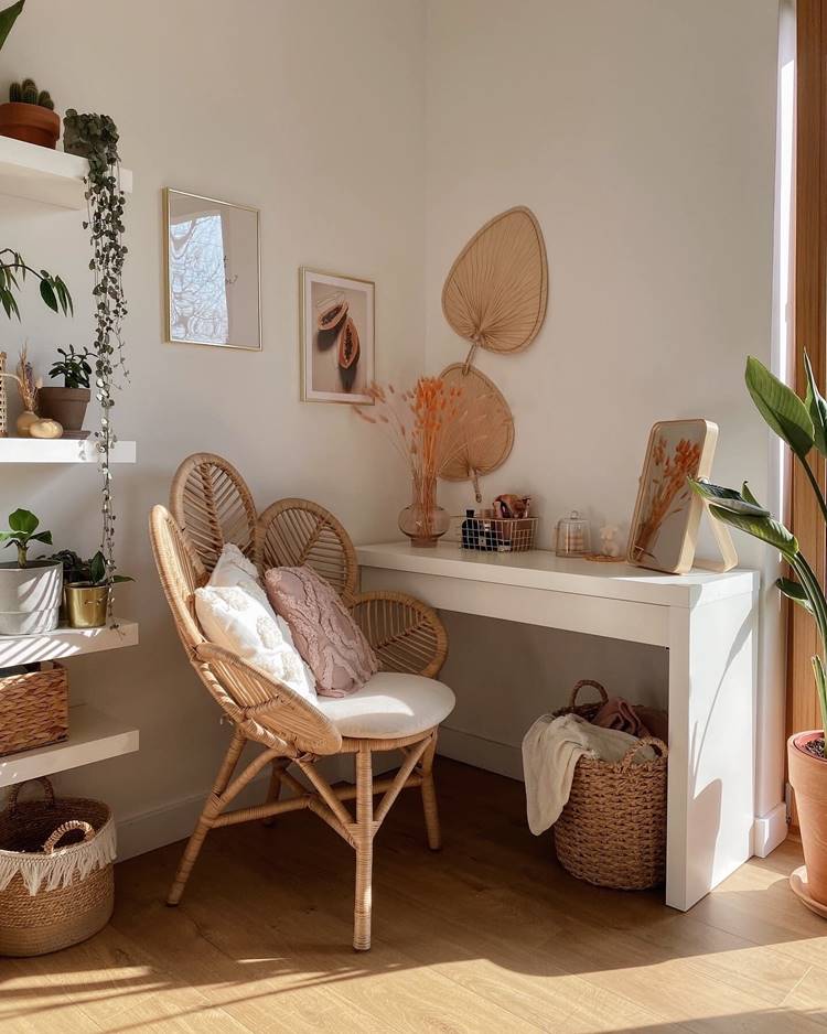 Summer decoration of a cozy corner of a room.  There is a wicker chair with white and light pink cushions, positioned next to a white table.  On the wall above the table hang two paintings and a leaf-shaped decorative object.  A variety of plants are present in the image;  some are on shelves mounted on the wall to the left, others on the table and on the floor.  The white table contains decorative objects, including a small vase with dried orange flowers.  A rectangular mirror rests against the wall on the table.  Two woven baskets are on the floor;  one contains a folded blanket.  The environment is indoors, possibly a living room, bathed in natural light.