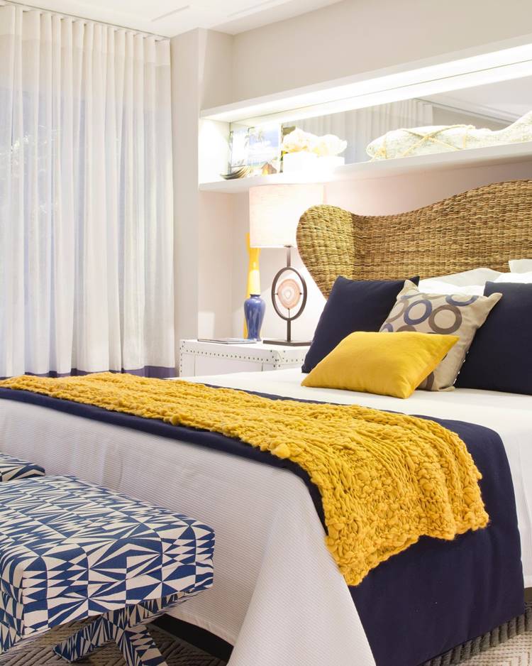 Summer decoration of an elegantly decorated room.  There is a large bed with white sheets, a dark blue blanket and a mustard yellow blanket.  Several pillows are on the bed, including two large dark blue pillows and a mustard yellow pillow.  A white nightstand sits next to the bed with a modern lamp.  Semi-sheer white curtains cover the window next to the bed, allowing natural light to enter the room.  Above the headboard are illuminated built-in shelves containing decorative items such as vases and boxes.  The room is bright and features a soft color palette.