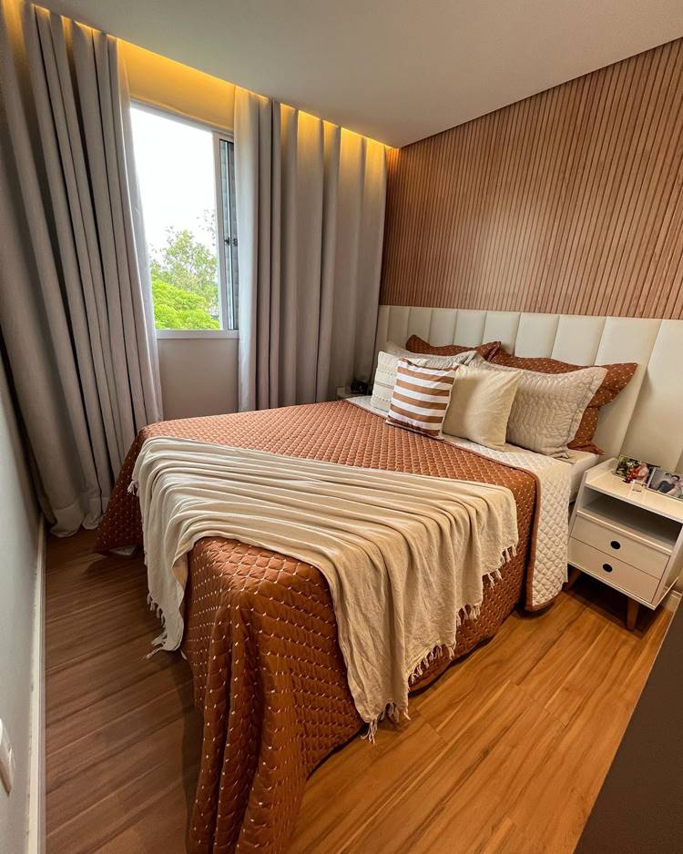 The image shows a bright room with a large bed.  The bed is covered in a textured brown bedspread and adorned with several decorative pillows.  The wall behind the bed has a textured design that complements the color and pattern of the bedspread.  There are dark gray curtains hanging in a window next to the bed, letting in natural light.  A white nightstand with drawers is located next to the bed.  The dark wood flooring adds a touch of elegance to the room.  The room is modern and cozy, with soft lighting.