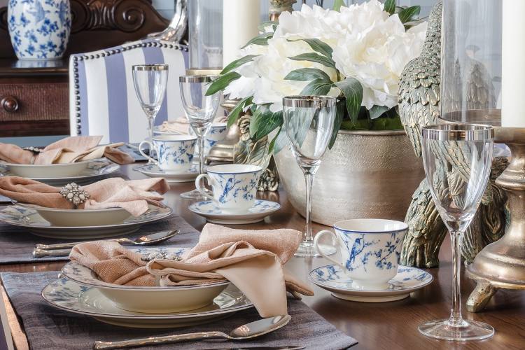 The image shows a wooden table. There are four white plates with the same floral pattern. The dishes are accompanied by silver cutlery and beige napkins. The table is decorated with a vase with white flowers and a figure of a bird. The table is served with crystal glasses and cups in the same pattern as the plates. The background consists of a blue and white cushioned wooden chair and a wooden sideboard with a blue and white vase. The image conveys a feeling of refinement and class.