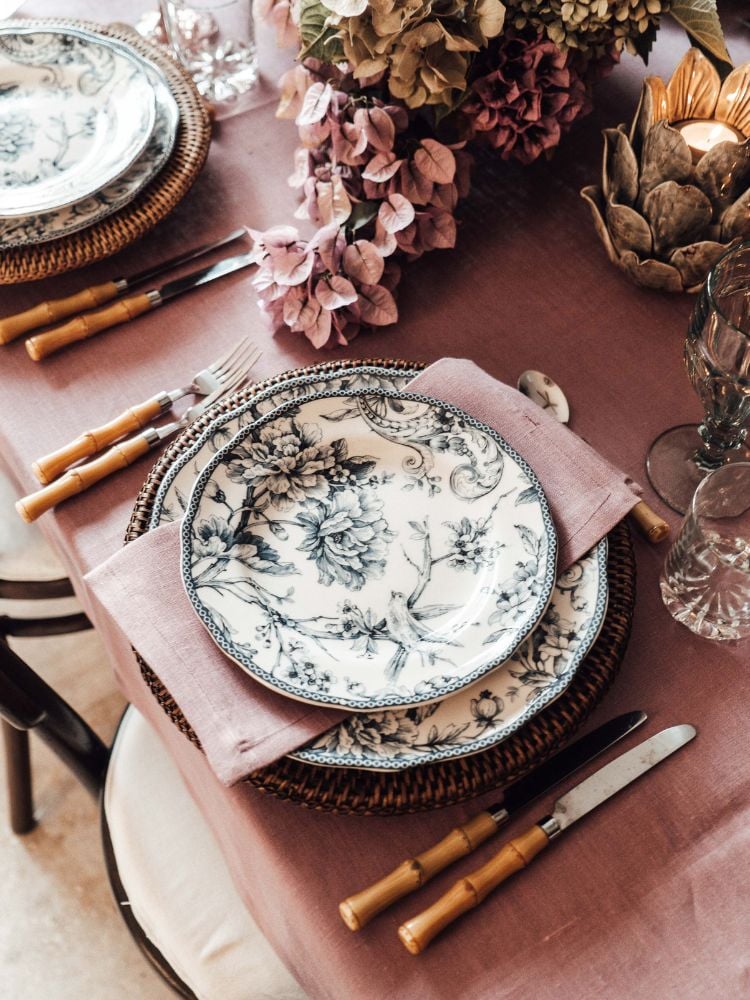 The image shows a table decorated with a floral and Portuguese theme.  The set table has a white plate with a black floral design, bamboo-handled cutlery and a pink napkin.  There are also pink flowers on the table.  The tablecloth is light pink. 