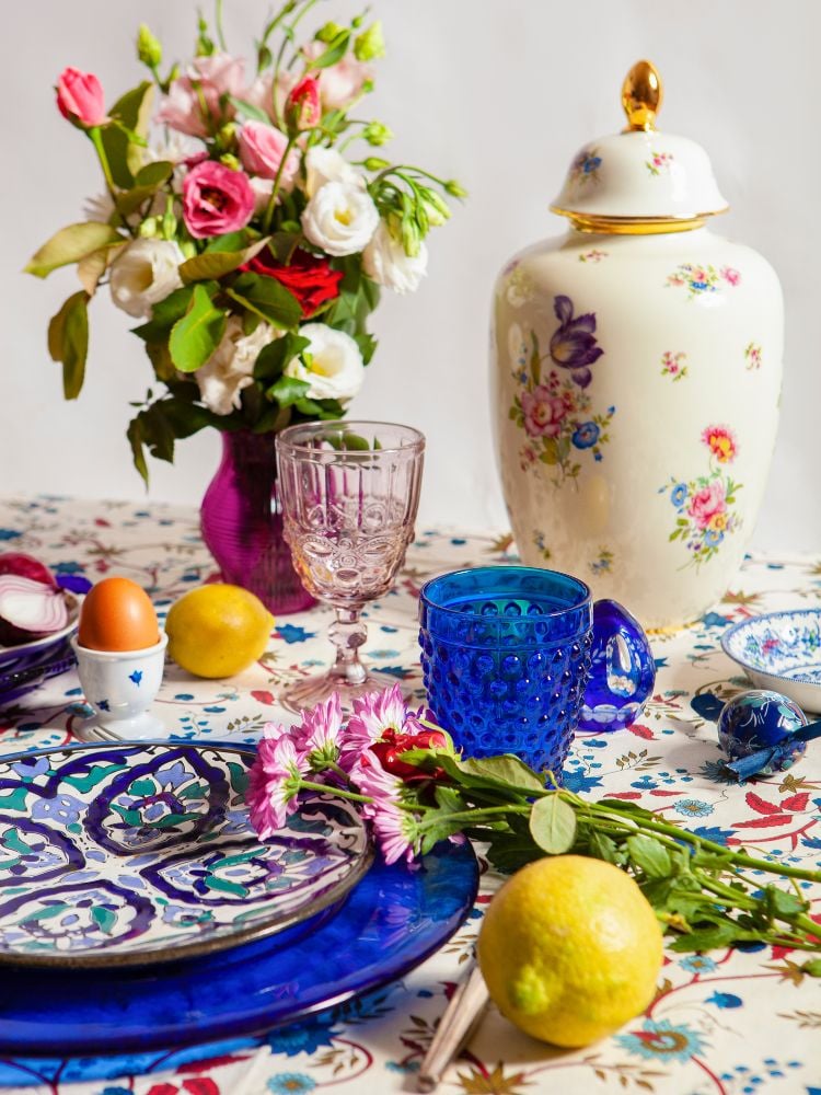 The image shows a table covered in a white cloth with a blue and red floral pattern.  The center of the table is a vase of red and white roses.  There is a white ceramic vase and a blue and pink floral design.  There are two glasses, one is a pinkish transparent wine glass and the other is a blue glass wine glass.  There is a blue and white ceramic plate with a pink flower on it.  There are two lemons and an egg on the table.  The background is a white wall.  The image conveys a feeling of freshness and romanticism.