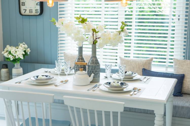 The image shows a white, rectangular table.  The table is set with white plates with blue floral patterns, white napkins and silver cutlery.  The center of the table is a vase with white flowers and a white teapot.  The background consists of a blue wall with a clock and a window with white shutters.  The background also includes a bench with blue and white cushions.  The image conveys a feeling of simplicity and comfort.