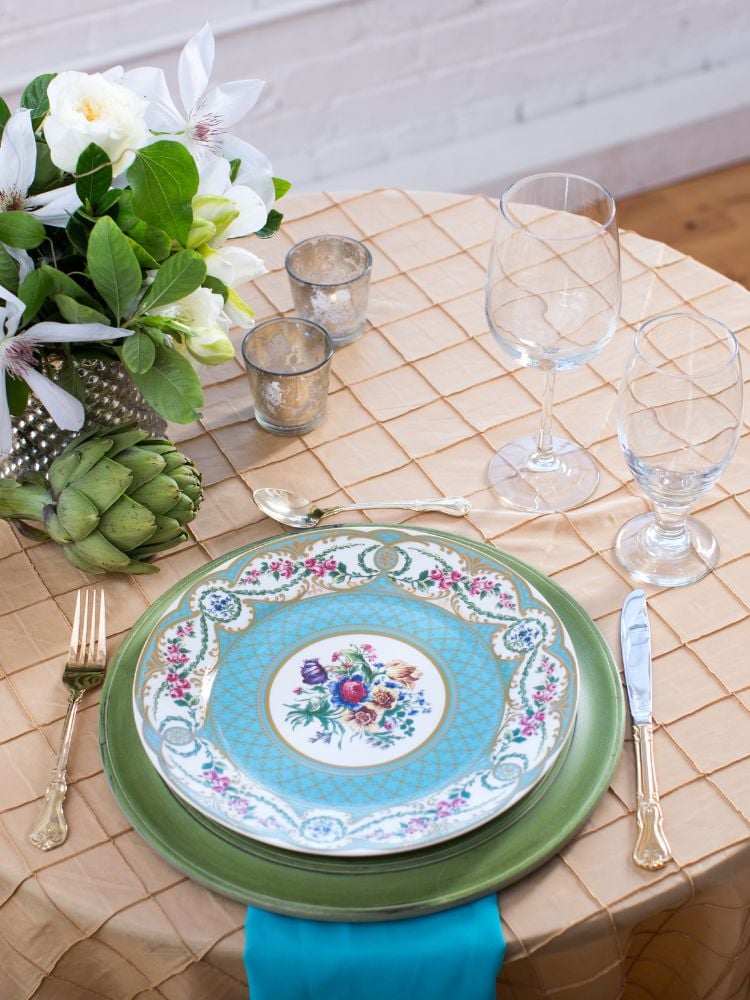 The image shows a table covered with a beige checkered cloth.  The table is set with a green plate below another plate with a blue and white floral design in the center.  The plate is on a blue napkin.  There are two glasses of wine on the table.  There is a vase of white flowers and a green artichoke on the table.  The table is served with a fork and knife with engraved handles.  The background is a white brick wall.  The image conveys a feeling of sophistication and good taste.