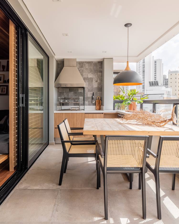 Balcony of the decorated 187 m² apartment.  Table with 4 chairs, with black and bronze lamp.  Decorating in the center, a dry rooted trunk.  In the background, a barbecue, alongside kitchen objects, such as a knife and cutting board