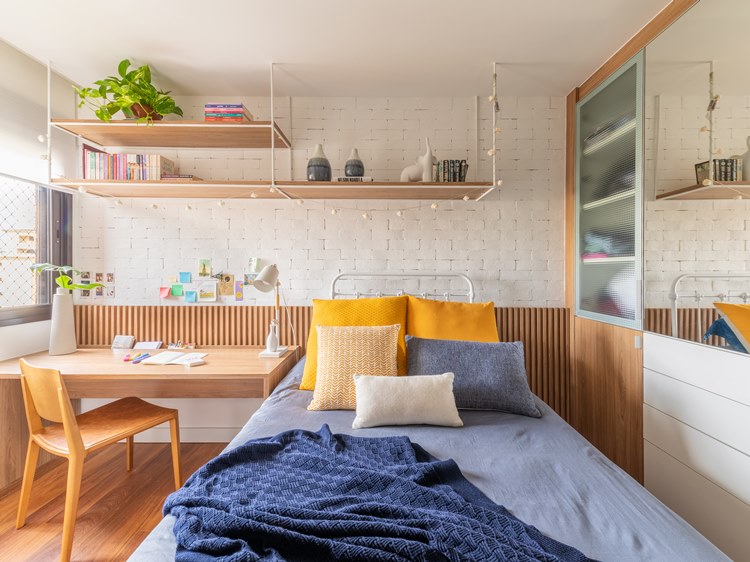 View of the wall from the headboard of the bed in the older sister's room.  Shelf above with books, objects and plant.  Next to the bed, a table with a chair for studies.