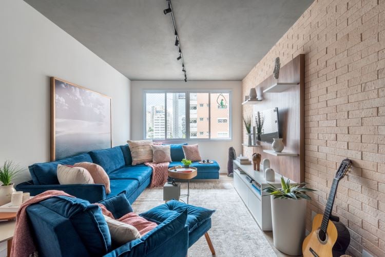 Living room with a large blue sofa, blue armchair, light pink cushions, television on the wall, painting behind the sofa and ample natural lighting coming from a huge window.