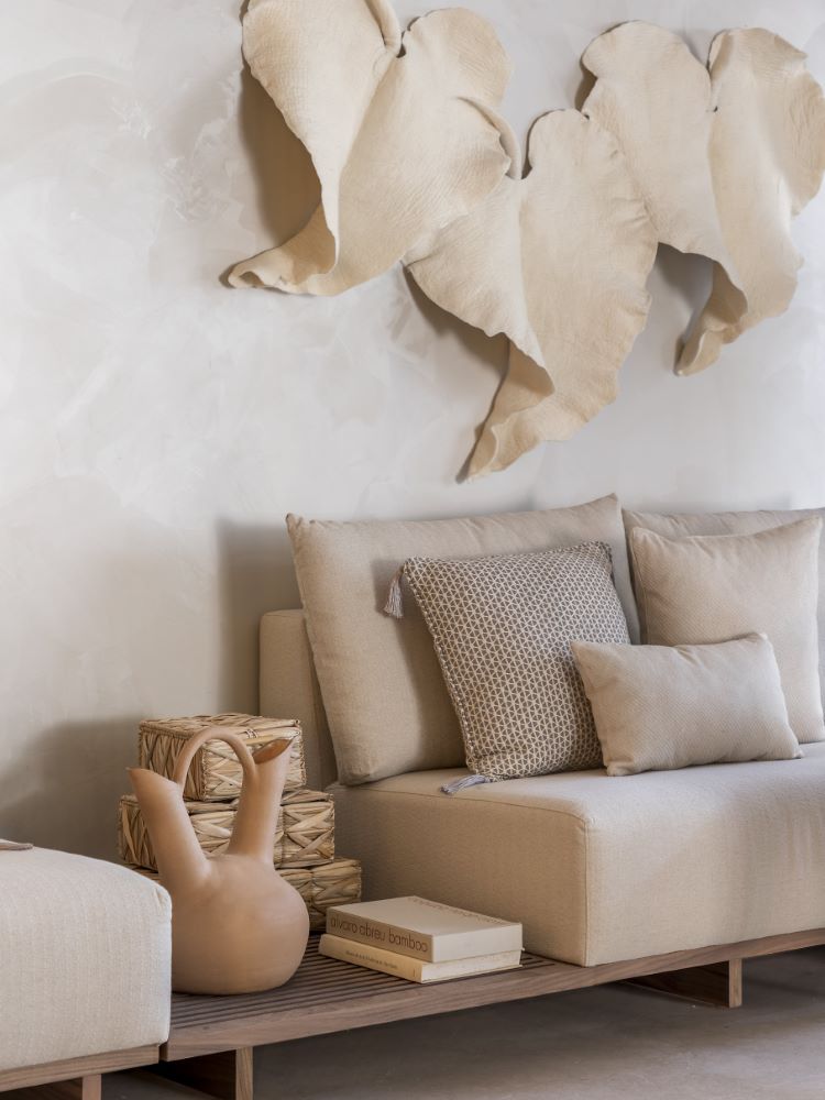 The image shows part of a beige sofa, with a ceramic vase next to it and handcrafted leaves on the wall above.