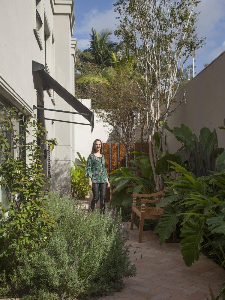 Image of an external area of ​​a house, with several plants and a light-skinned woman in the background, wearing black pants and a green blouse.