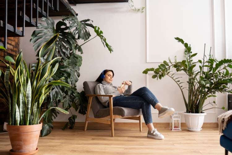 Light-skinned, dark-haired woman wearing headphones and holding a cell phone.  She is sitting in an armchair, arranged in a relaxing environment with plants and neutral colors, such as white and wood tone, below a black staircase.  Decoration contributes to mental health