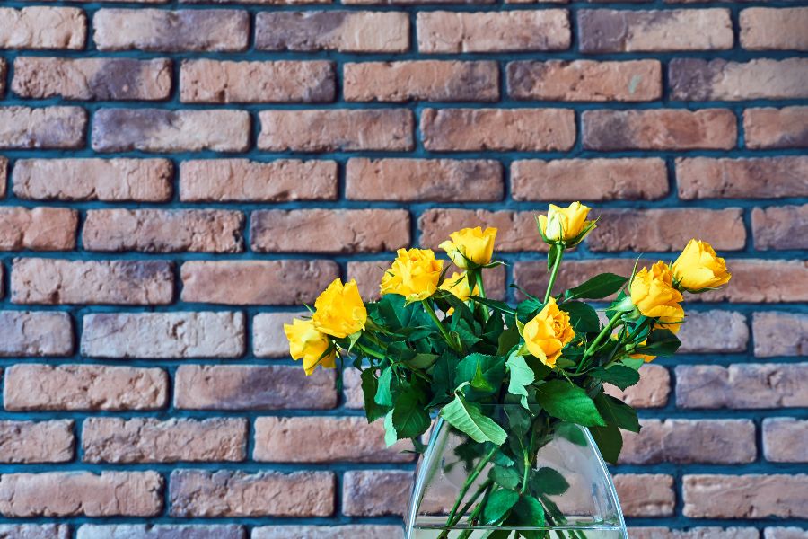 Glass vase with yellow roses.  In the background there is a brick wall