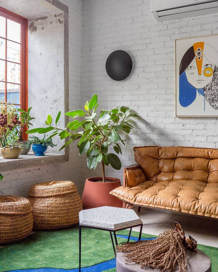 The image shows a cozy and bright living room with summer decor.  There is a brown leather sofa.  In front of the sofa, there are two round organizer boxes made of natural braided material.  A small side table with a marble top is next to the sofa.  A lush green plant sits in a terracotta pot on the floor, adding a touch of nature to the room.  Other smaller plants are positioned in the window.  On the wall next to the sofa is a colorful artwork depicting a person's stylized face.  The room has white brick walls that give it a rustic, modern feel, and a large window with a red frame that lets in lots of natural light.  The decor has a summery touch, with bright colors and plants that bring a bit of nature into the home.