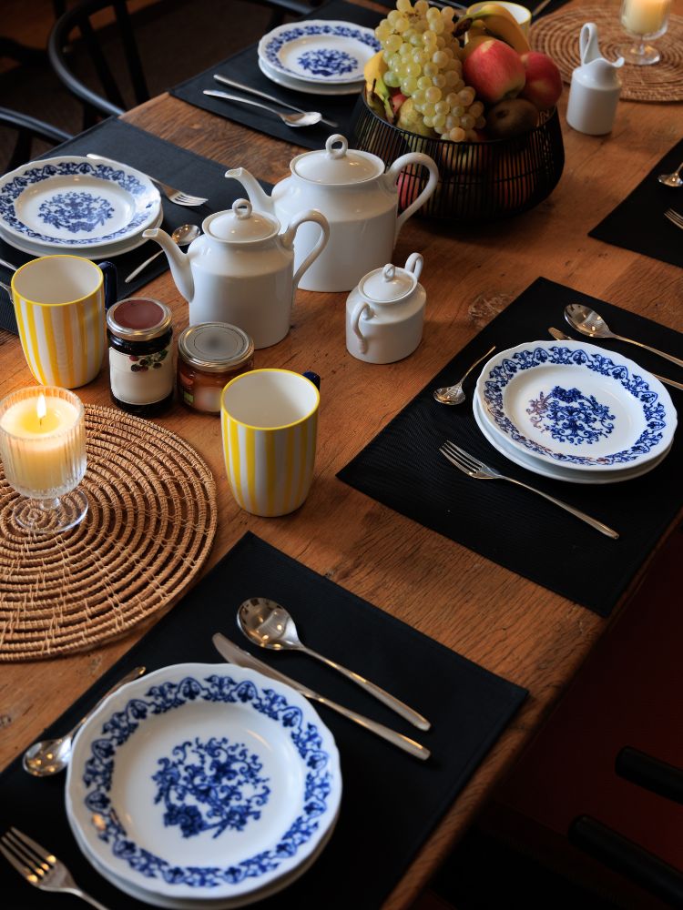 Table set in Portuguese style with a wooden table with a black cloth covering it.  There are four white plates with blue floral designs, one on each corner of the table.  Each plate has a silver spoon next to it.  There are also four straw placemats under the plates.  In the center of the table, there is a fruit basket with grapes and apples.  There is also a white teapot and sugar bowl, and two yellow and white striped candles.  The image conveys a feeling of warmth and tradition. 