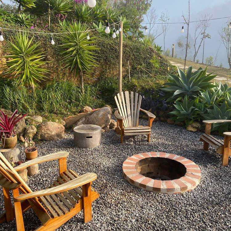 Jardim com lareira de pedra, cadeiras de madeira e decoração com plantas ao redor, além de varal de luzes e cerca de toras de madeira com casinhas de passarinhos no chão. Há grama e alecrim plantado em volta da cerca.