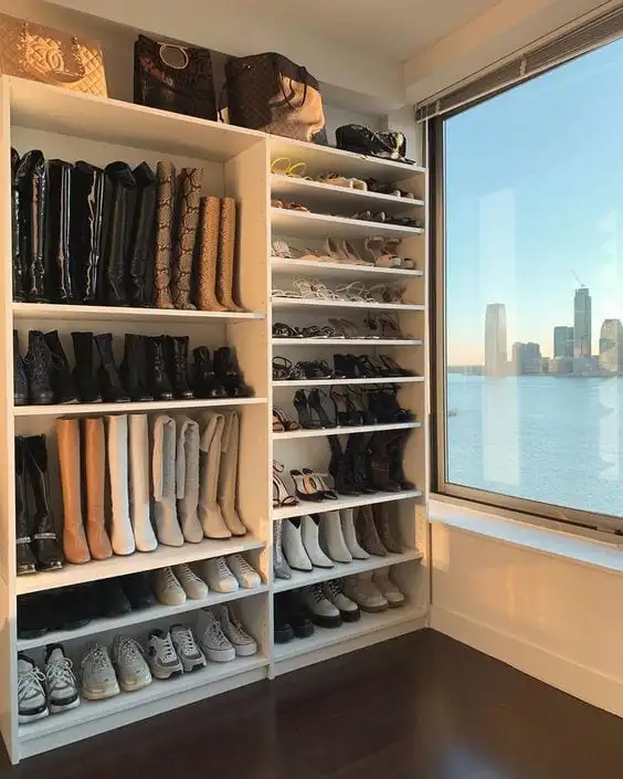 Closet com janela com vista para um grande rio com prédios ao fundo. A sapateira está repleta de tênis, botas, saltos e coturnos.