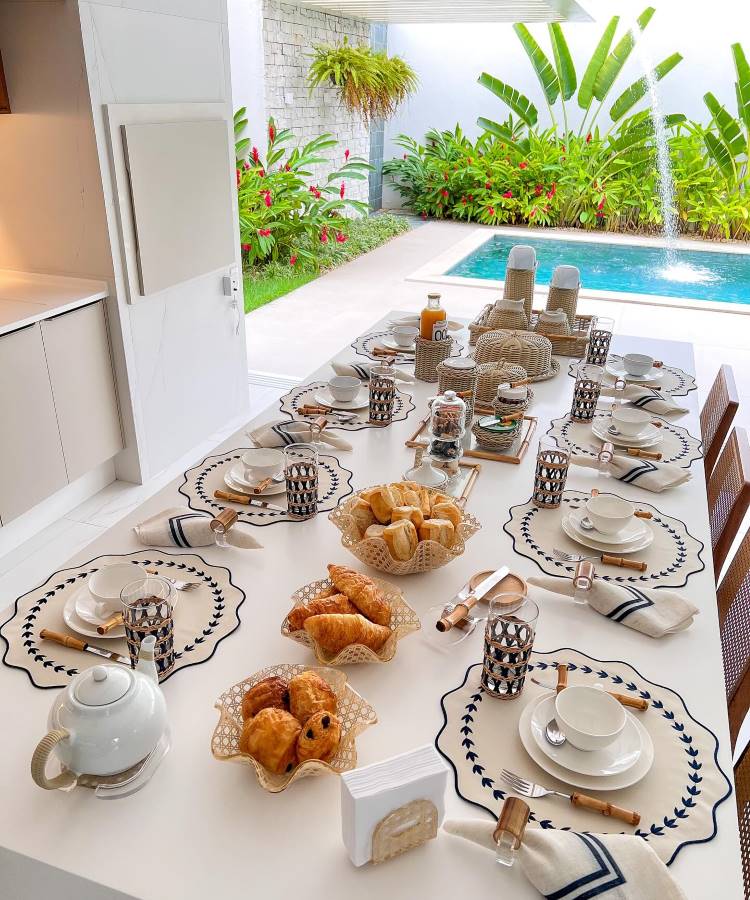 The image shows a table set for breakfast in an outdoor setting, next to a swimming pool. There are sousplats with blue decorative borders, white plates and matching cups. Cutlery with bamboo handles and fabric napkins add elegance. In the center, baskets of bread and croissants are arranged next to a tray with juice and jars of jam. Glasses with geometric details complete the storage, creating an inviting and sophisticated environment. In the background, a tropical garden adds freshness to the scene.