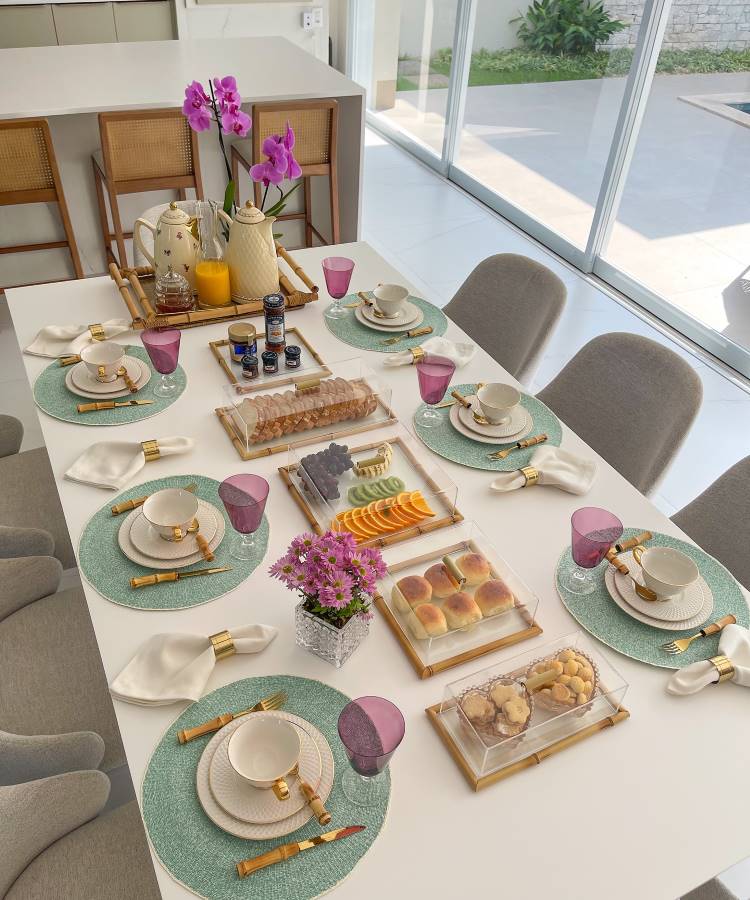 The image shows a table set for breakfast in a bright environment. There are green sousplats under white plates and cups with gold details. Bamboo cutlery and white napkins with gold rings complement the arrangement. Purple cups add a pop of color. In the center, trays contain bread, cookies, cold cuts and sliced ​​fruit. A jug of juice and jars of jam are arranged next to a coffee maker. A vase with purple flowers decorates the table, creating a welcoming and elegant atmosphere.