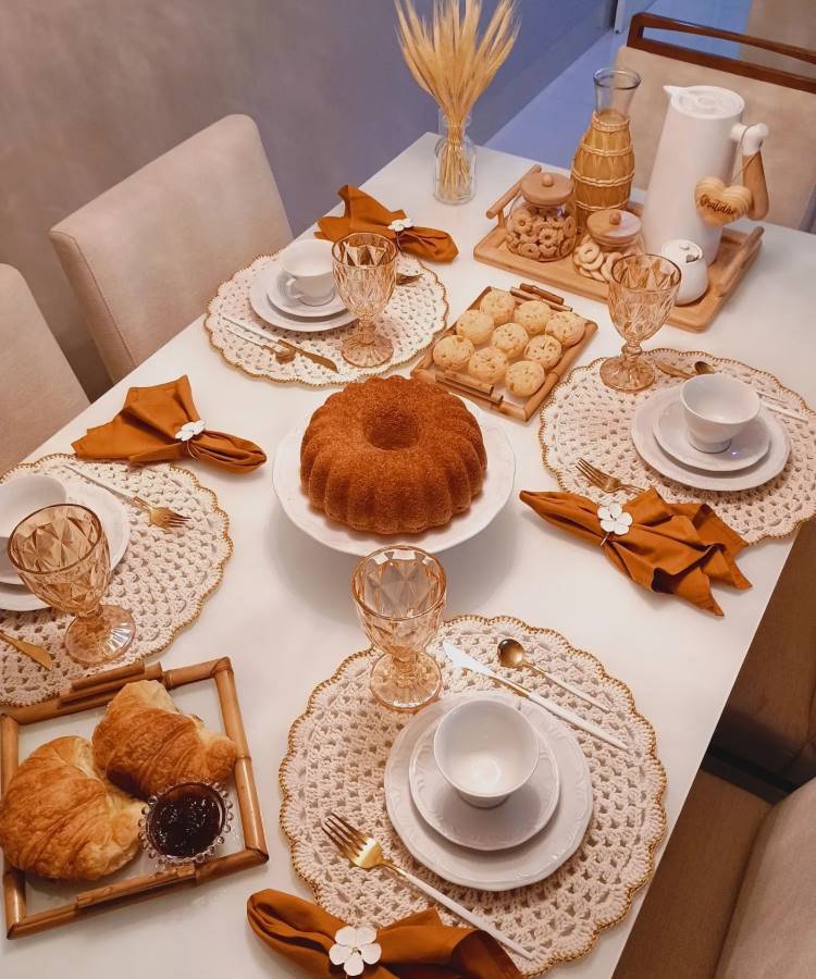 The image shows a table set for breakfast, with beige crochet sousplats and white plates. The golden cutlery is accompanied by orange napkins with floral details. In the center, a round cake is placed on a white plate. Around there are croissants with jam, biscuits and cheese bread on wooden trays. Glass cups with golden details and a white thermos complete the scene, creating a welcoming and harmonious environment.