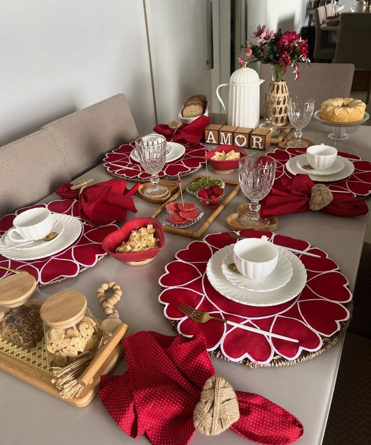 The image shows a table set for breakfast, highlighting red heart-shaped sousplats. White plates and cups are accompanied by red napkins with straw napkin holders. Elegant glass bowls complete the storage. In the center, there is a white thermos and wooden blocks forming the word "AMOR". Wooden trays hold cookies and bread, while an elevated plate displays a cake. A vase with red flowers adds charm to the room, creating a romantic and welcoming setting.