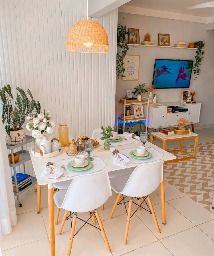 The image shows a well-lit and warmly decorated dining room. There is a white rectangular table with wooden legs, surrounded by four white chairs with wooden legs. On the table, there are white dishes with green details, glass cups and folded napkins. In the center, a vase with a green plant adds a touch of freshness. Next to the table, there is a cart on wheels, which contains a jug of orange juice, cups and an arrangement of white flowers. A straw pendant lamp illuminates the table. In the background, there is a white bookcase with shelves, decorated with plants, paintings and decorative objects. A television is on, showing an image of a hummingbird. The floor has a geometric pattern in neutral tones, and there is an overall feeling of harmony and comfort in the room.