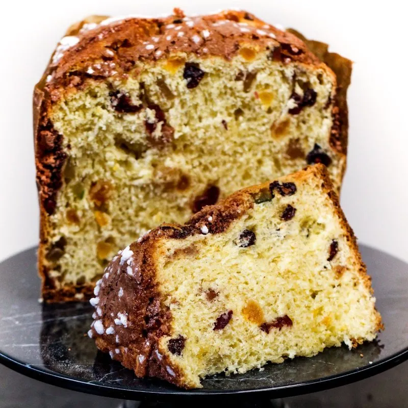 Panetone tradicional de frutas cortado ao meio mostrando o miolo e uma fatia sobre um pedestal.