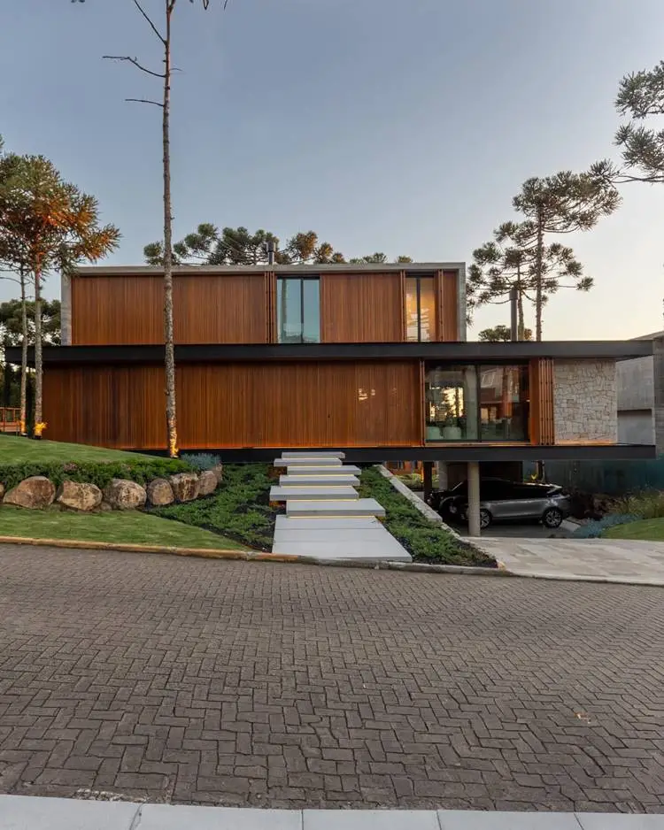 The following image shows a casa moderna, with walls made of wood and concrete, with large windows and is surrounded by trees. The house has a full staircase to white, which leads to the entrance, a parking garage, opened the car, and with a clear sky and so on