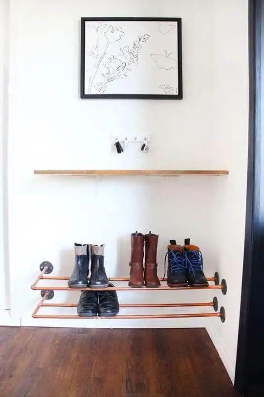 Chaussure rack faite de tuyaux dans les tons de cuivre, fixée sur un mur blanc. Dans ce qui précède, il est une tablette avec une porte, une clé et un tableau blanc. 