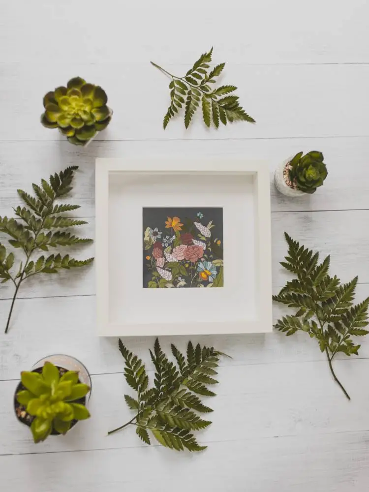 Imagem de um quadro de flores com moldura branca rodeado por folhagens e vasos de suculenta. 