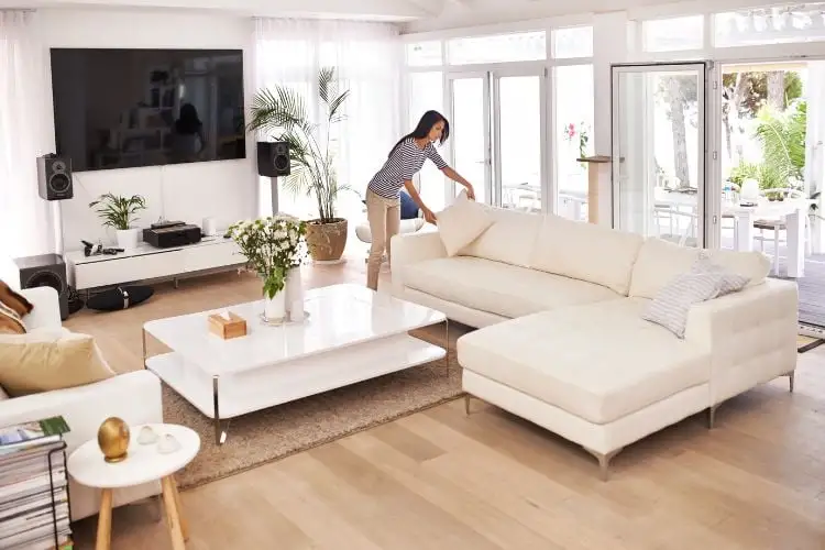 Woman, light skinned, packing up the cushion of the sofa, the beige in the living room with wooden floors, a TV and large windows